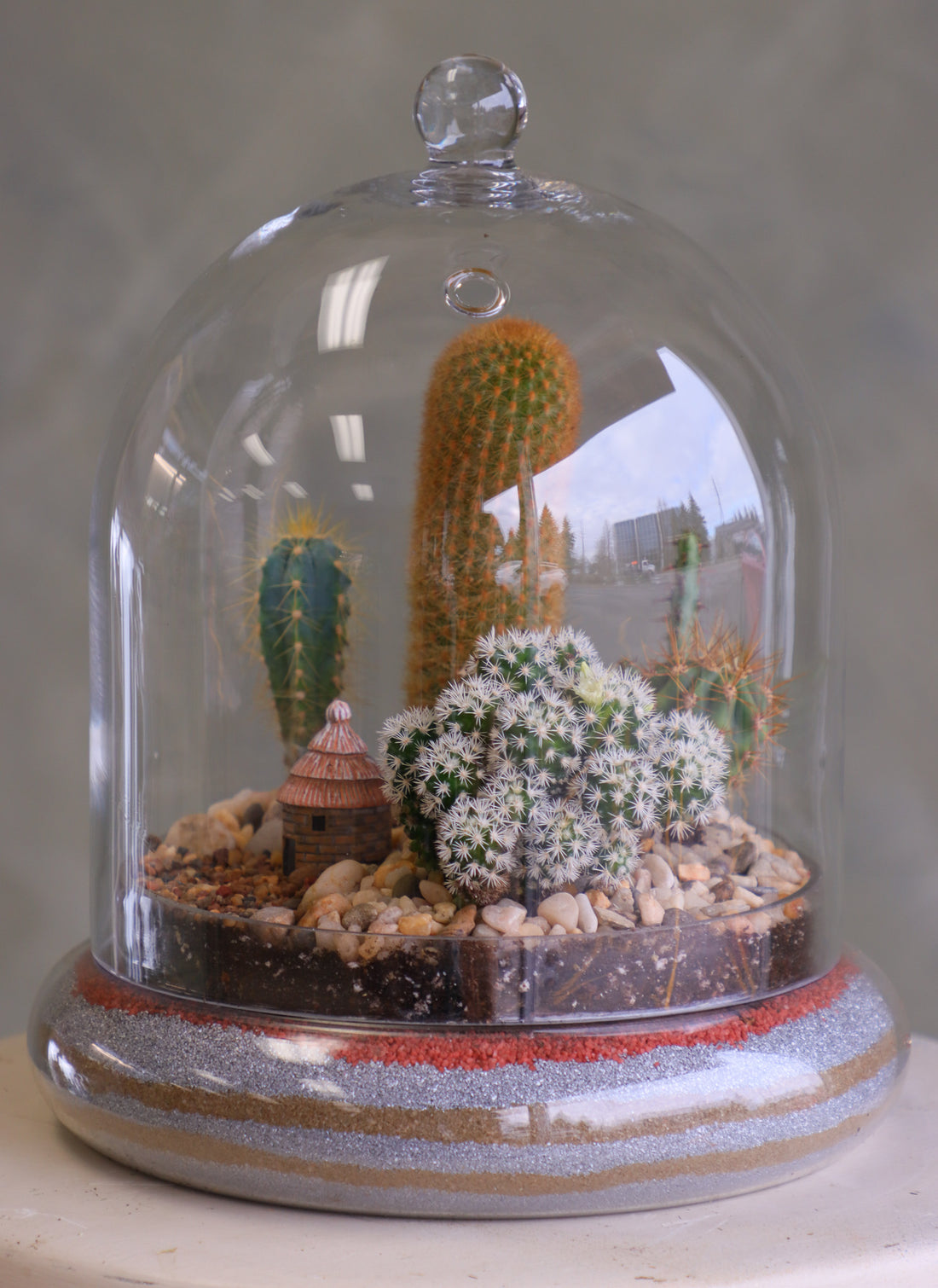 Glass cloche terrarium with mixed cacti, pebbles, layered sand base, and a tiny clay hut, styled as a miniature desert village on a round pedestal.